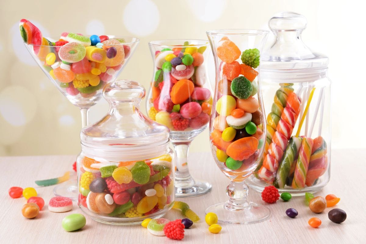 Several glass jars and glasses filled with assorted colorful candies and sweets are displayed on a light wooden surface.