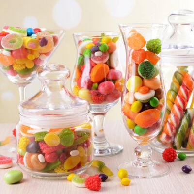 Assorted colorful candies displayed in glass jars and glasses on a light wooden surface with a soft, blurred background.