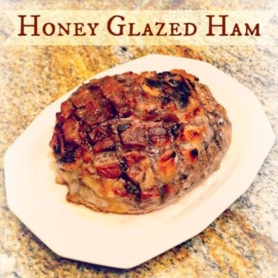 A honey glazed ham with a caramelized, scored surface sits on a white platter atop a granite countertop—perfect for adding to your collection of Easter recipes.