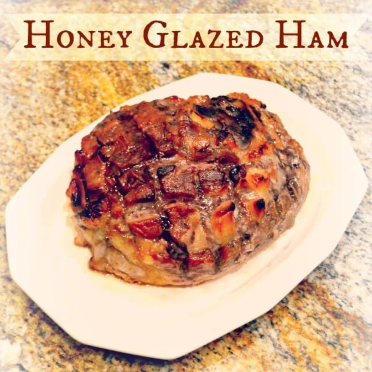 A honey glazed ham with a caramelized, scored surface sits on a white platter atop a granite countertop—perfect for adding to your collection of Easter recipes.