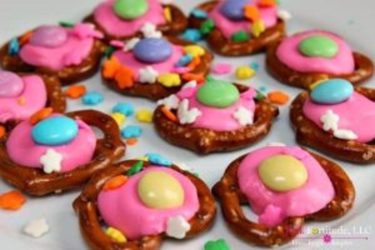 Mini pretzels topped with melted pink candy, colorful chocolate buttons, and assorted sprinkles make a fun addition to your collection of Easter recipes, all beautifully arranged on a white surface.