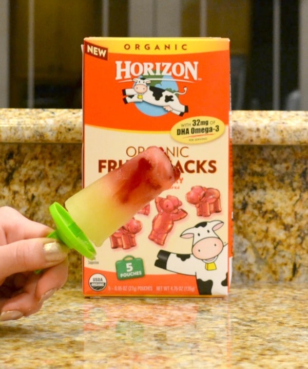 a hand holding a popsicle made out of fruit snacks and apple juice in front of a box of fruit snacks on a kitchen counter
