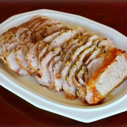 Herb roasted pork loin, sliced and arranged on a white oval platter, reveals golden brown edges and a juicy interior.