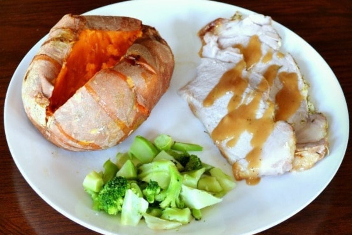 A plate with slices of roasted pork loin topped with gravy, a baked sweet potato, a serving of steamed broccoli.