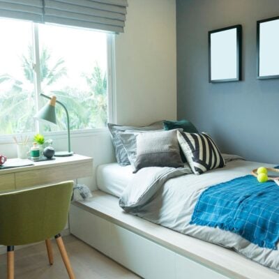 Organize and Decorate Your Kid’s Bedroom with These Simple Steps: A neatly made bed with pillows and a blue throw sits next to a desk and chair by the window; two blank frames hang on the gray wall.