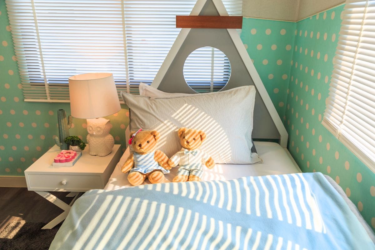 A neatly made bed with two teddy bears sits next to a nightstand with a lamp and decor, showing how to Organize and Decorate Your Kid's Bedroom with These Simple Steps in a polka-dotted, light-filled children's room.