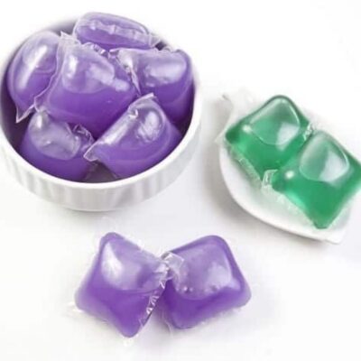 Several purple and two green laundry detergent pods displayed in small white bowls on a white surface, highlighting how, like other uses for dishwasher pods, these colorful capsules can offer versatile cleaning solutions.