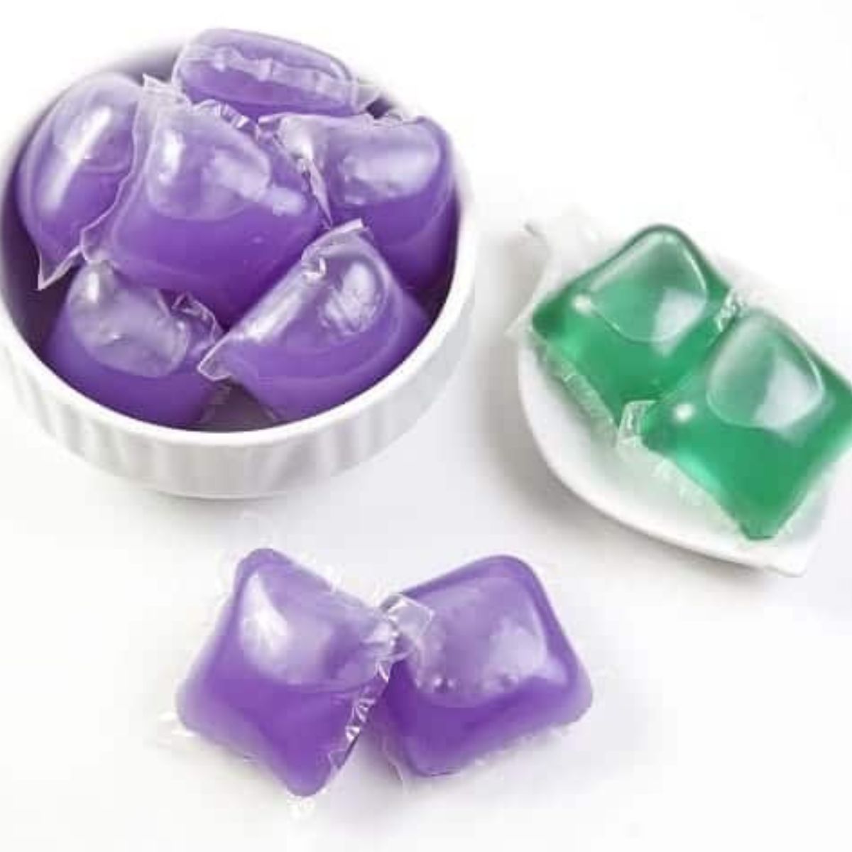 Several purple and two green laundry detergent pods displayed in small white bowls on a white surface, highlighting how, like other uses for dishwasher pods, these colorful capsules can offer versatile cleaning solutions.