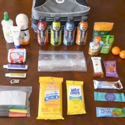 A collection of travel essentials on a table, including drinks, snacks, toiletries, disinfecting wipes, a first aid kit, and ziplock bags—just what you’d find in a well-organized sports mom bag—are neatly arranged in rows near a travel bag.
