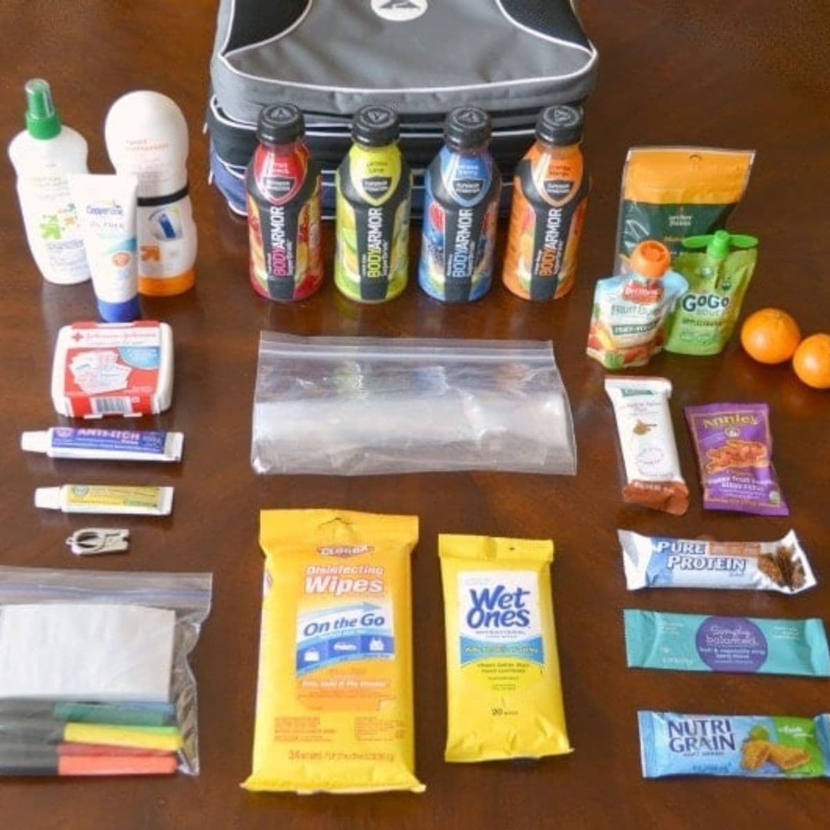 A collection of travel essentials on a table, including drinks, snacks, toiletries, disinfecting wipes, a first aid kit, and ziplock bags—just what you’d find in a well-organized sports mom bag—are neatly arranged in rows near a travel bag.