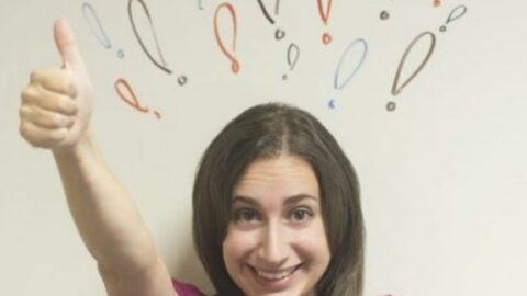 Woman smiling and giving a thumbs up, with colorful exclamation and question marks drawn on the wall behind her head—showcasing how positive gestures can improve your mood.