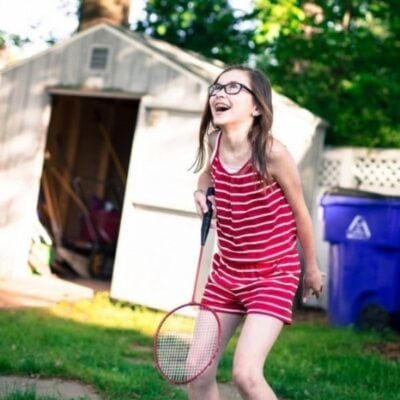 A young girl in a red striped outfit holds a badminton racket and smiles outdoors near a shed and recycling bin, ready to show that games you can play with your kids instead of exercising can be just as fun and active.