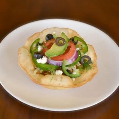A chicken tostada topped with sliced tomato, red onion, avocado, jalapeño, black olives, and crumbled cheese on a white plate, set against a brown background.