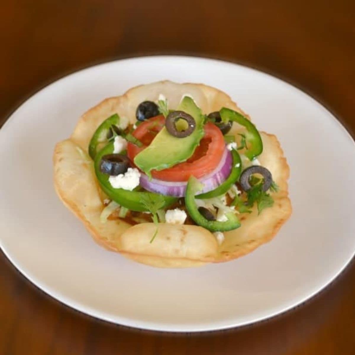 A chicken tostada topped with sliced tomato, red onion, avocado, jalapeño, black olives, and crumbled cheese on a white plate, set against a brown background.