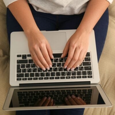A person types on a laptop keyboard while sitting on a beige couch, viewed from above, focused on a blog post stockpile challenge.