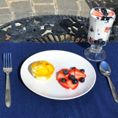 White plate with breakfast casserole cup, strawberries, and blueberries. Glass dish with fruit parfait. Fork and spoon. All on blue place mat