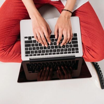 Person in red pants sits cross-legged, typing on a laptop atop their lap—perhaps working on a blog post stockpile challenge—viewed from above.