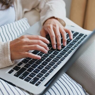 Close-up of a person typing on a laptop, sitting with a striped pillow on their lap, possibly planning their next blog post or taking part in a blog post stockpile challenge.