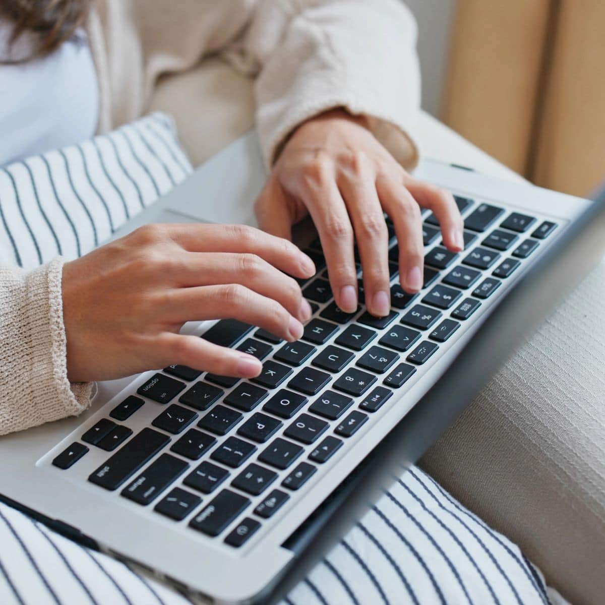 Close-up of a person typing on a laptop, sitting with a striped pillow on their lap, possibly planning their next blog post or taking part in a blog post stockpile challenge.