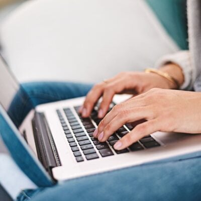 Close-up of a person typing on a laptop keyboard, seated on a couch and wearing a bracelet and jeans.