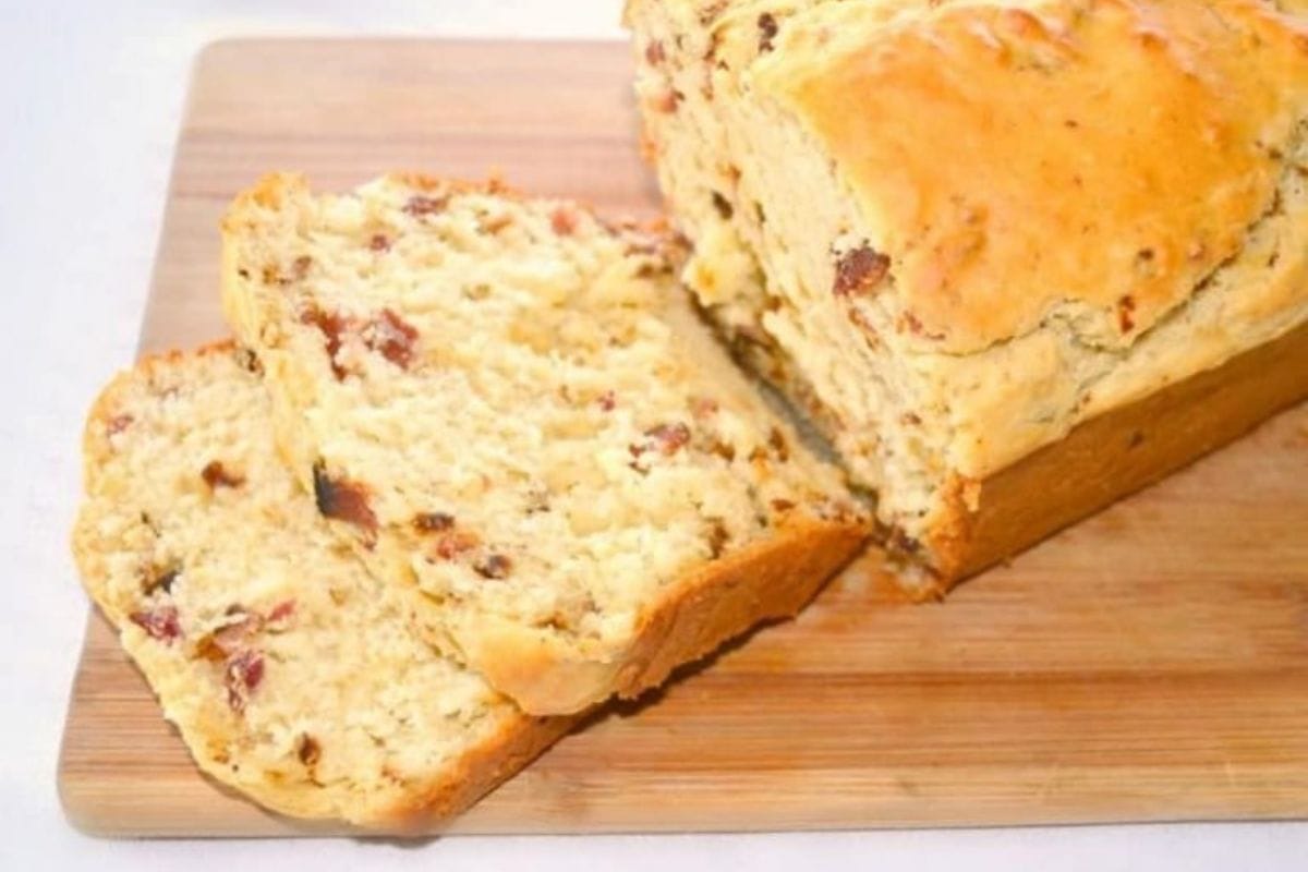 A loaf of bacon bread with two slices cut, on a wooden cutting board.