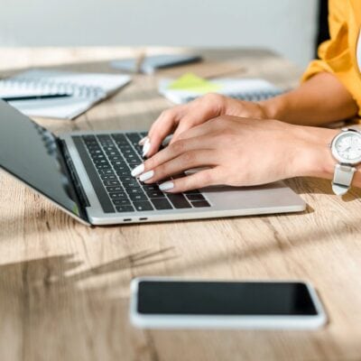 Close-up of a person typing on a laptop at a wooden desk, with a smartphone, notepad, and watch visible.