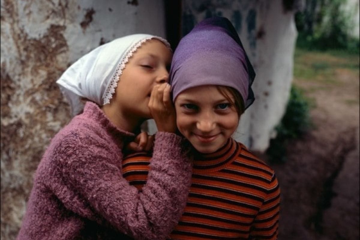 Two young girls wearing headscarves stand close together, one whispering into the other's ear-a scene that hints at childhood secrets and reminds us why you should not confide in your child. The girl being whispered to smiles at the camera.