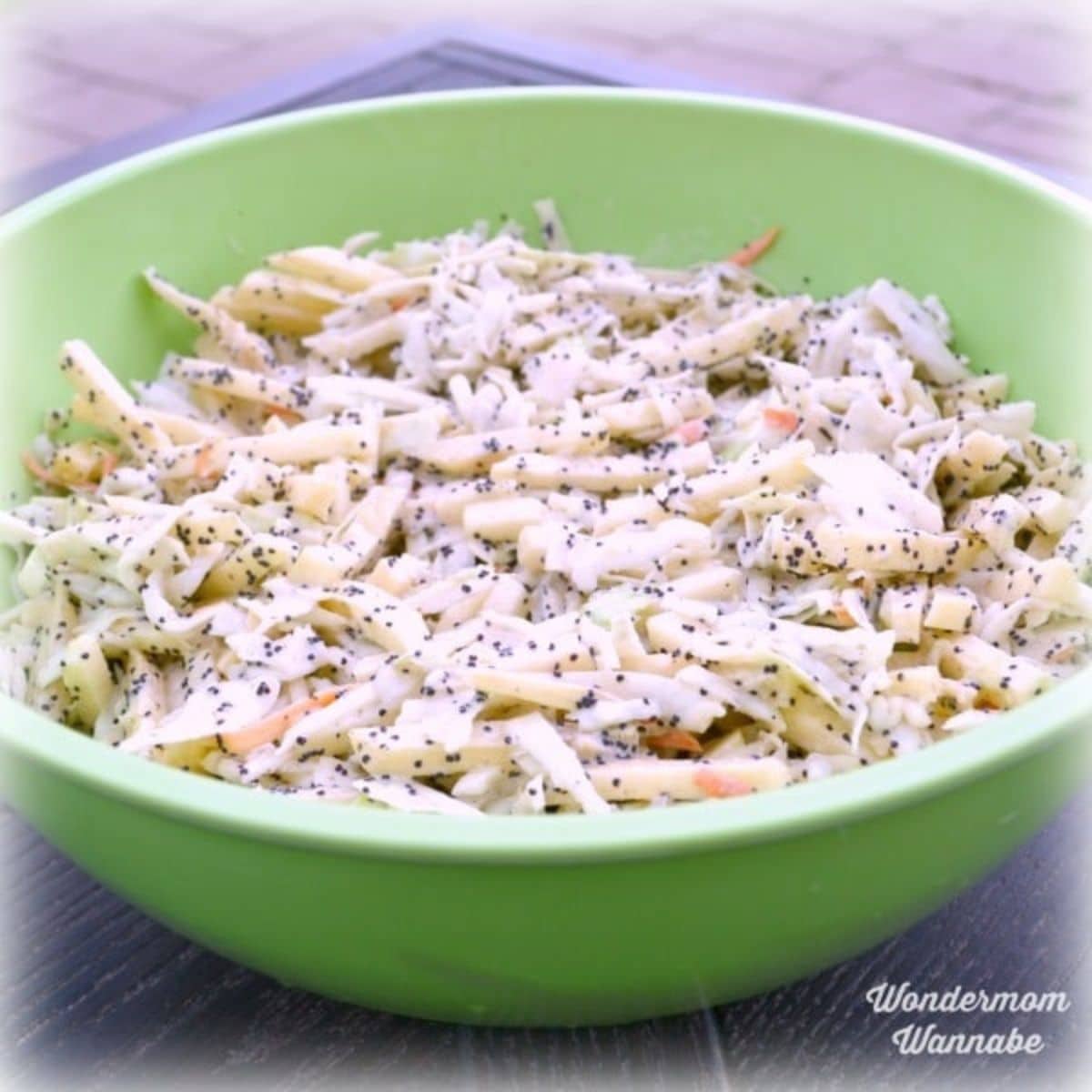 A green bowl filled with shredded apple coleslaw salad topped with poppy seeds and grated cheese.