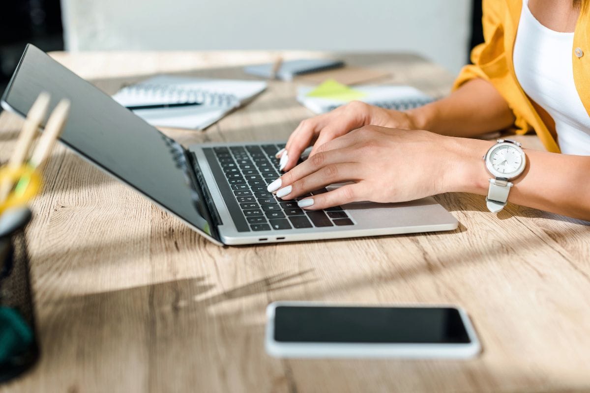 Close-up of a person typing on a laptop at a wooden desk with a smartphone, notebooks, and pencils nearby.