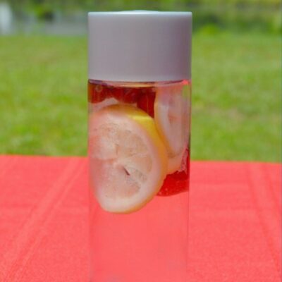 A clear bottle of cranberry lemon infused water with lemon slices inside, placed on a red surface and green grass in the background.