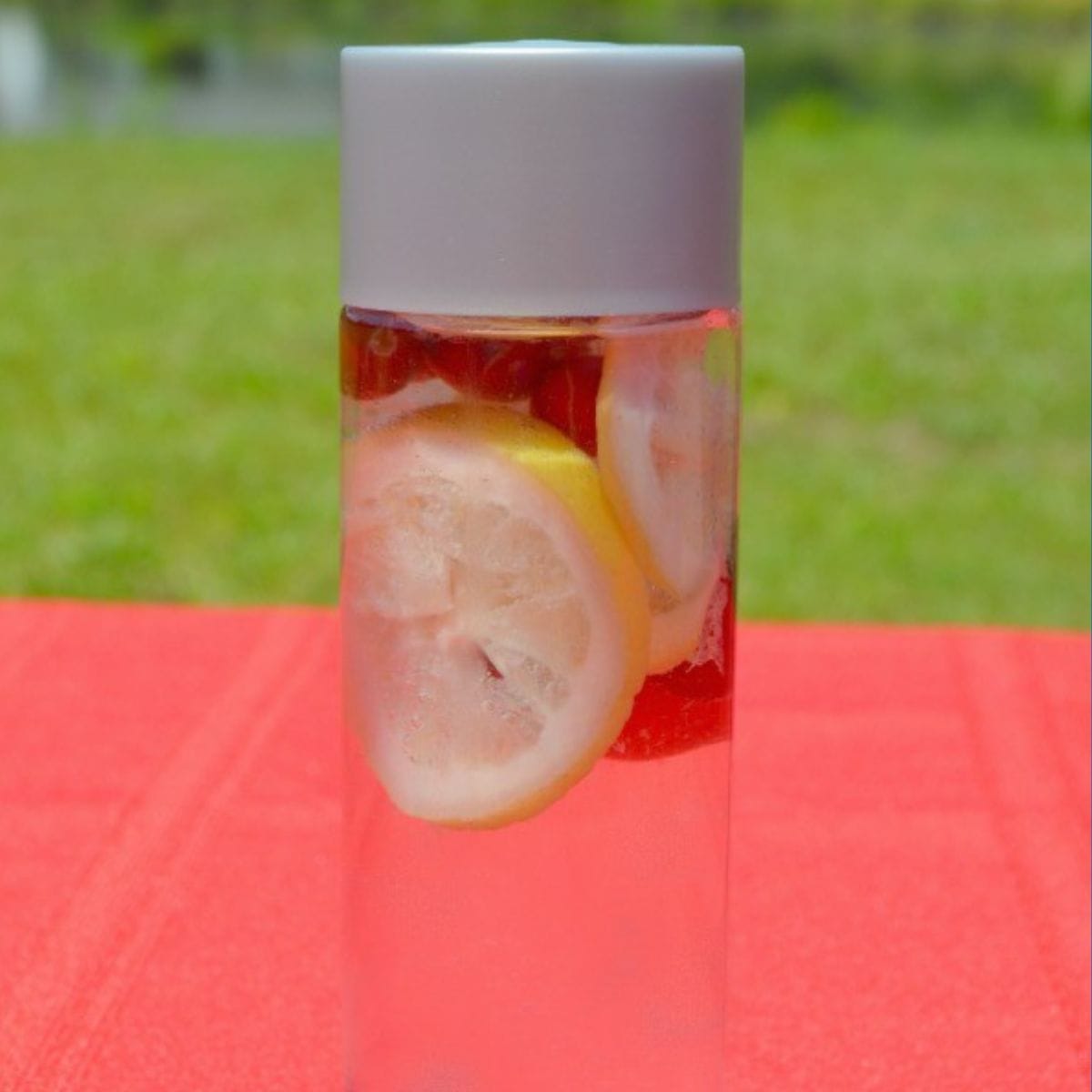 A clear bottle of cranberry lemon infused water with lemon slices inside, placed on a red surface and green grass in the background.