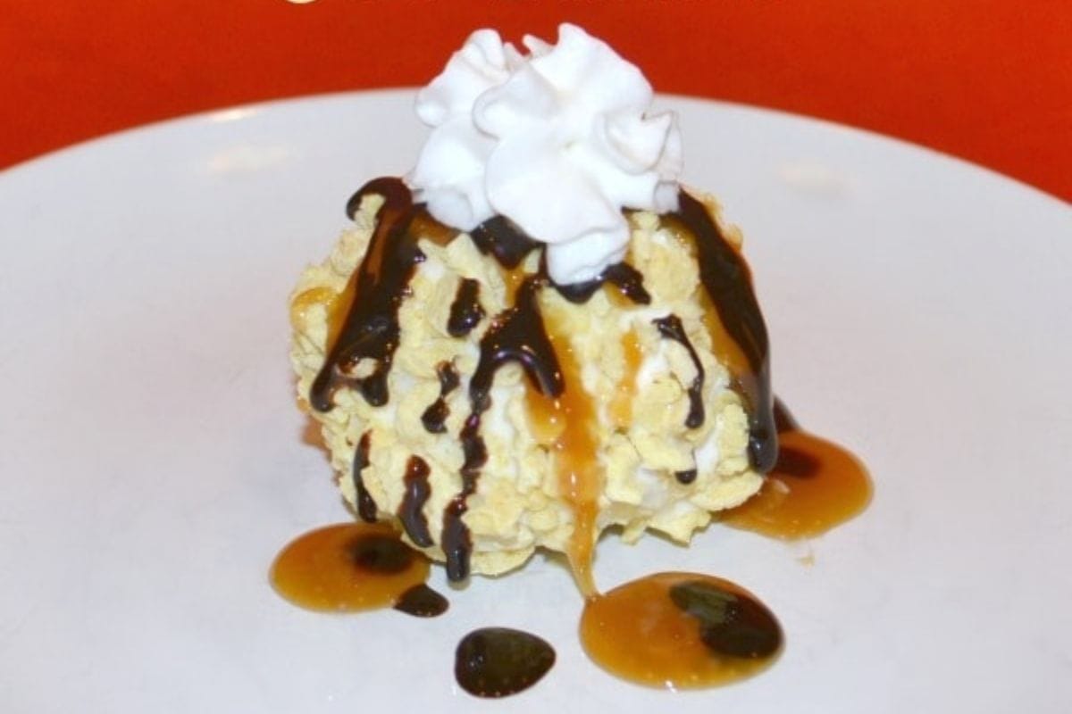 A scoop of crunchy unfried ice cream topped with whipped cream, drizzled with chocolate and caramel sauce, served on a white plate with an orange background.