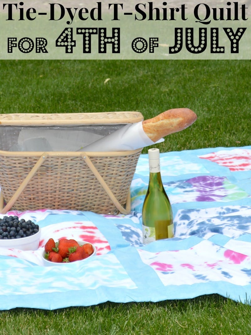 a picnic basket, bread, fruit and a bottle on a picnic blanket on the grass with title text reading Tie-Dyed T-Shirt Quilt For 4th of July