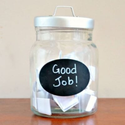 A glass reward jar with a metal lid is labeled "Good Job!" and contains several folded pieces of paper inside.