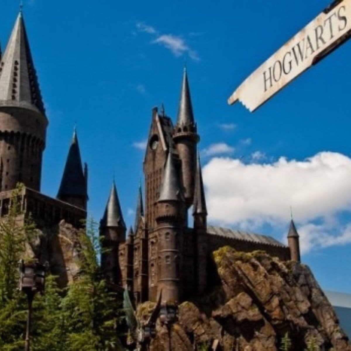 A stone castle with tall spires sits on a rocky hill under a blue sky. A sign reading "Hogwarts" points toward the castle.