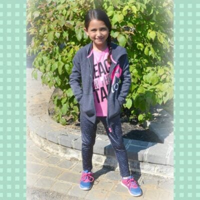 A girl stands outdoors on a paved path, showcasing back to school kid fashion in a grey jacket, pink shirt, dark pants, and sneakers, with green foliage in the background.