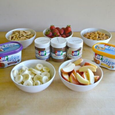 Bowls of sliced bananas, apple wedges, strawberries, cookies, and crackers surround various Jif spreads—whipped and salted caramel flavors—perfect for an end of summer snackation on a wooden surface.