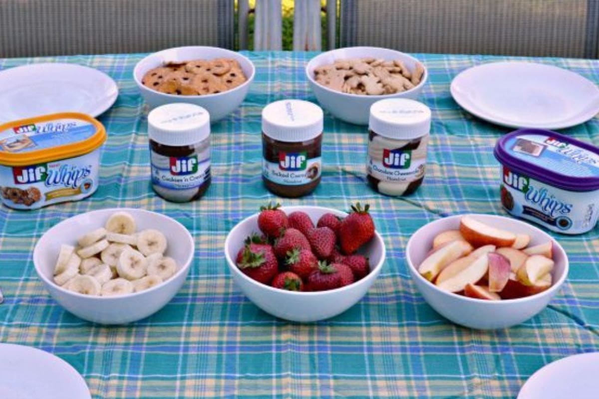 A table with bowls of banana slices, strawberries, apple slices, cookies, and Jif spreads on a blue plaid tablecloth.