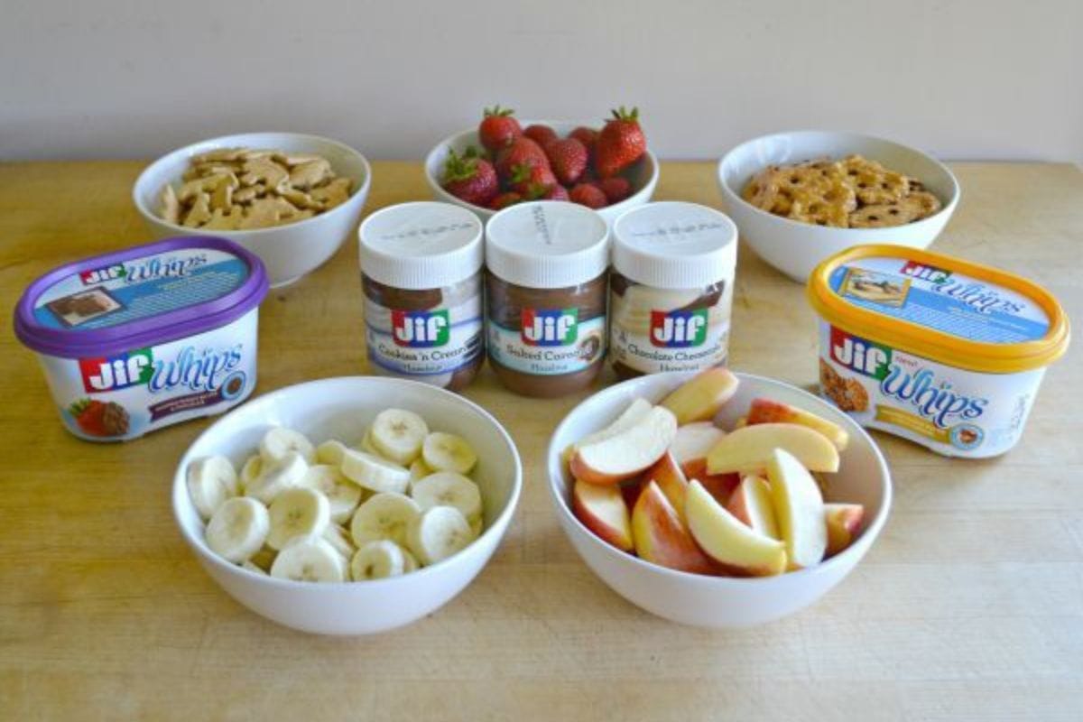 Fruit and crackers in white bowls and jif whips and spreads on a brown table as end of summer Snackation Snacks.