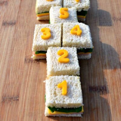 Six square sandwiches arranged in a hopscotch pattern on a wooden surface, each topped with a yellow number from 1 to 5, create playful sandwich art.