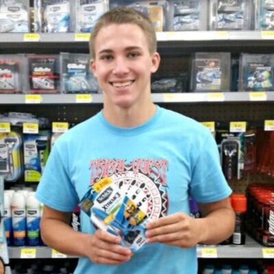 A young man in a blue t-shirt smiles while holding a pack of Schick Xtreme3 razors, shopping for dorm living in front of a store shelf filled with shaving products.