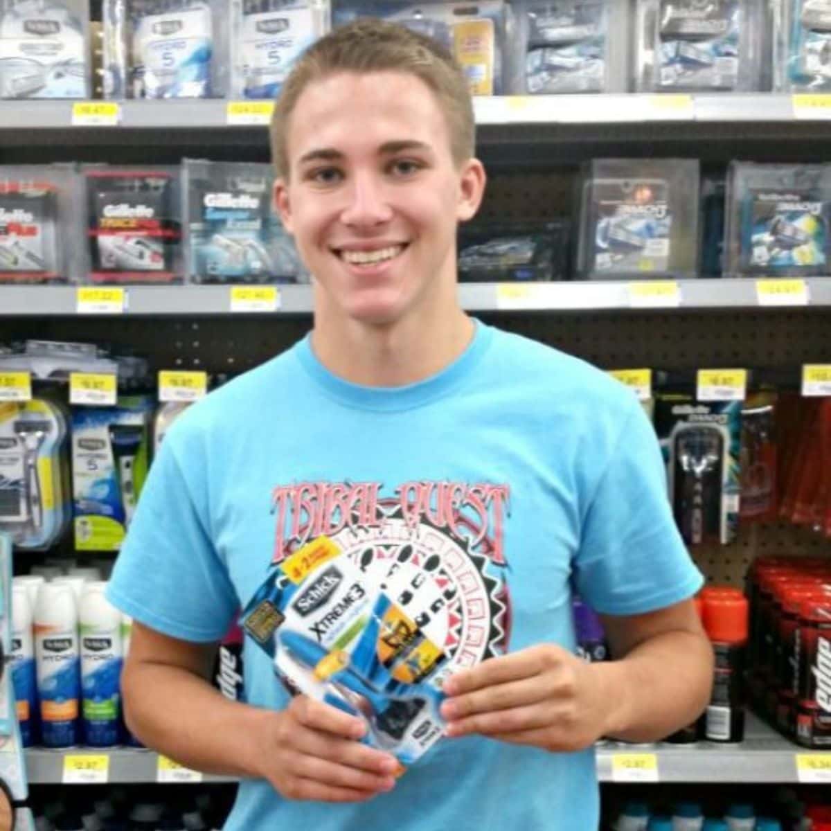 A young man in a blue t-shirt smiles while holding a pack of Schick Xtreme3 razors, shopping for dorm living in front of a store shelf filled with shaving products.