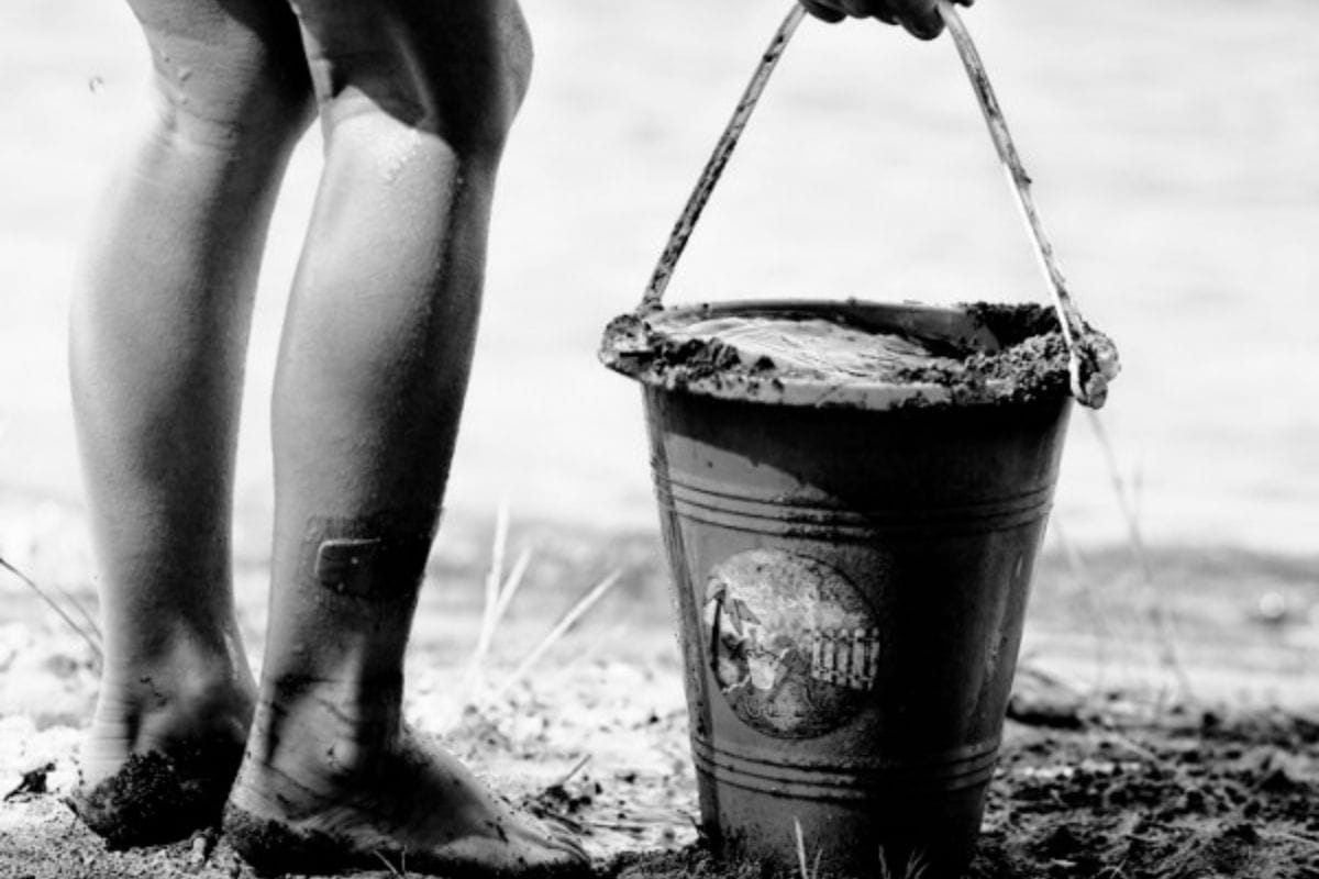 Black and white close-up of a child's legs and feet covered in sand and mud, standing next to a bucket filled with water and mud-capturing simple moments that are among the best tips for getting through the summer with kids.