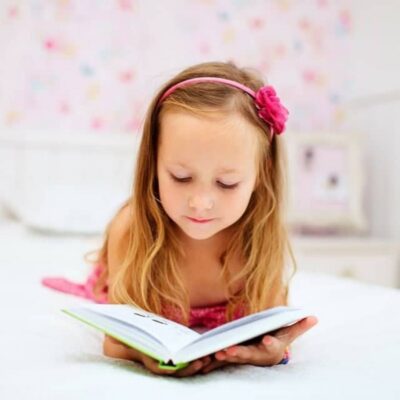 Young girl with long blonde hair and a pink headband lies on her stomach on a bed, showcasing her reading ability as she enjoys a book in a brightly lit room.