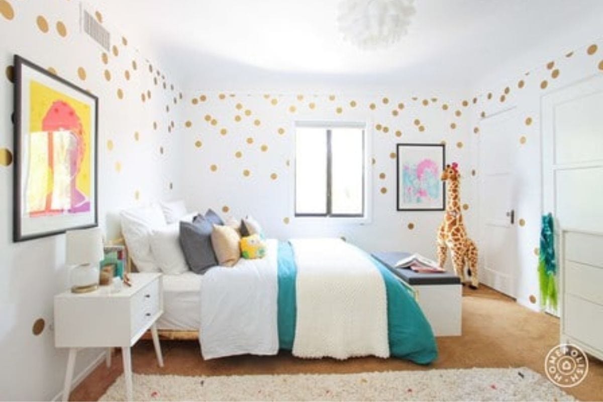 Bright bedroom with white walls and gold polka dots, a bed with teal and white bedding, framed art, a large stuffed giraffe, and a white nightstand-perfect inspiration for Kids' Room Decorating Ideas.