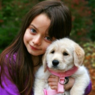 A young girl with long brown hair holds a light-colored puppy in a pink harness, both looking at the camera outdoors—perfect inspiration for those seeking first pets for small kids.