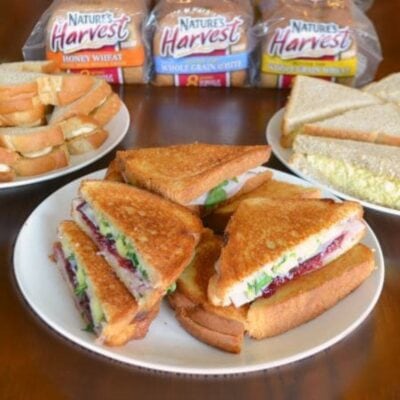 A plate of grilled sandwiches with turkey, lettuce, and cranberry sauce sits in front of two plates showcasing sandwich varieties for the whole family, alongside packs of Nature's Harvest bread.