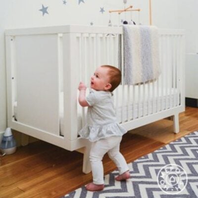A baby stands holding the side of a white crib in a nursery with a gray and white rug, striped blanket, and star wall decals—perfect inspiration if you’re looking for tips for decorating a nursery.