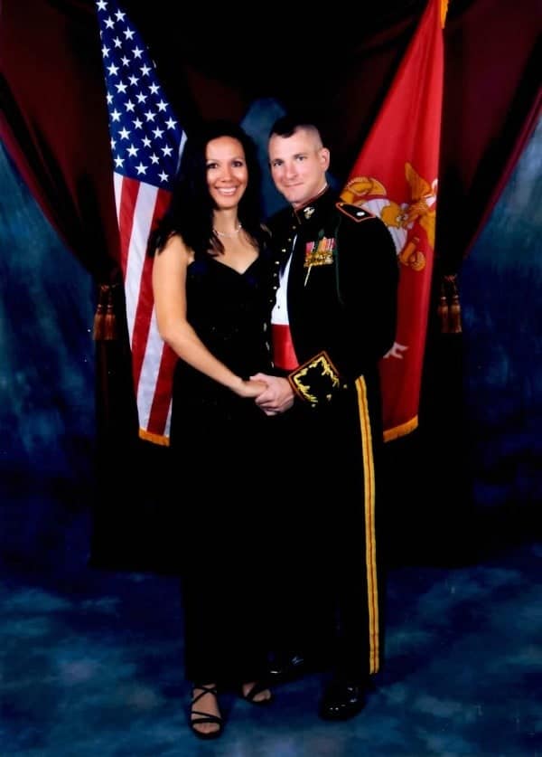 a wife and her husband dressed in his Marine uniform standing in front of an American flag and the Marines flag