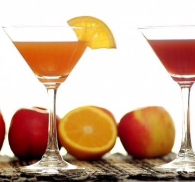 An orange cocktail and a red cocktail with oranges and apples in the background